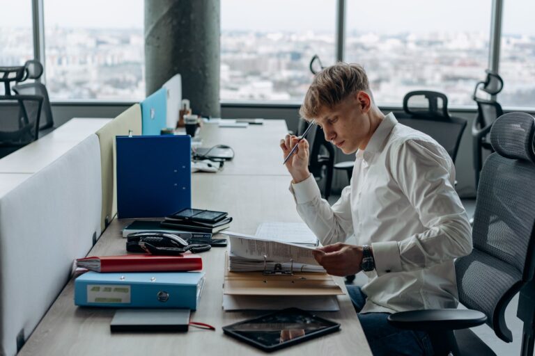 Man in white shirt concentrating on paperwork at modern office desk with city view.