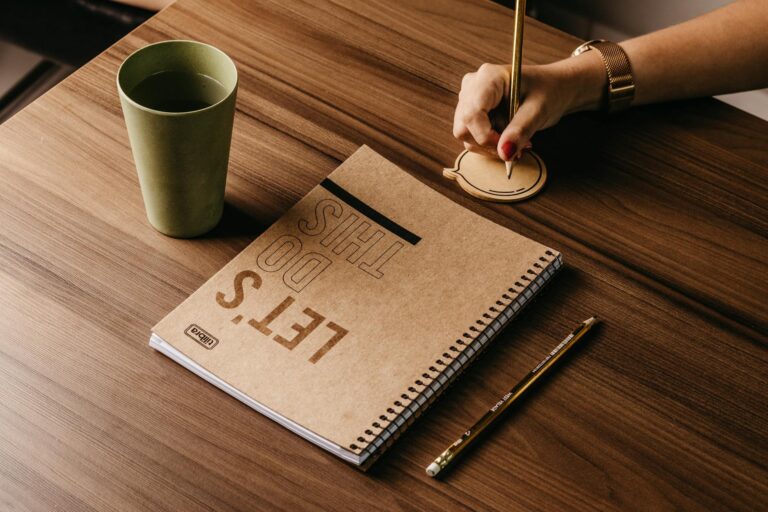 A woman's hand writing on a speech bubble notepad next to a cup and notebook on a wooden desk.