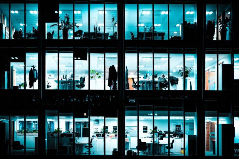 Night view of an illuminated modern office building showcasing workspaces through large windows.