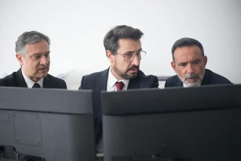 Team of three businessmen in suits and glasses collaborating at desks, focused on shared screens.